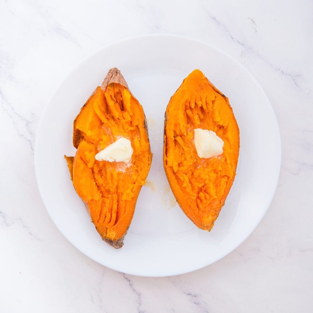 A white plate holds a baked sweet potato split in half, each half topped with a small pat of butter.