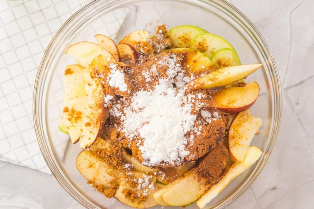 A clear glass bowl filled with sliced red and green apples, topped with brown sugar, cinnamon, and a heap of white powdered starch, on a light marble surface next to a checkered cloth.