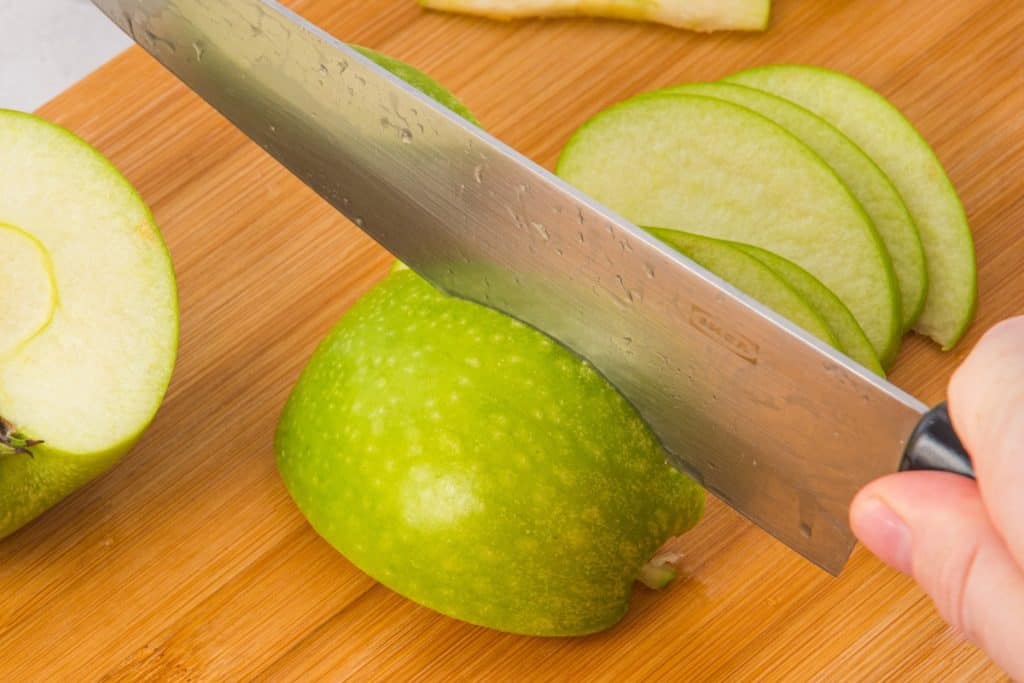 A hand holds a knife slicing a green apple on a wooden cutting board, with several thin apple slices and another halved apple visible nearby.