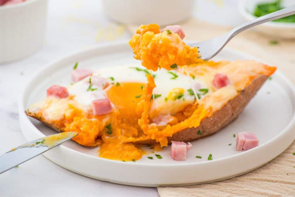 A baked sweet potato topped with a runny egg, diced ham, and chopped herbs is served on a white plate.