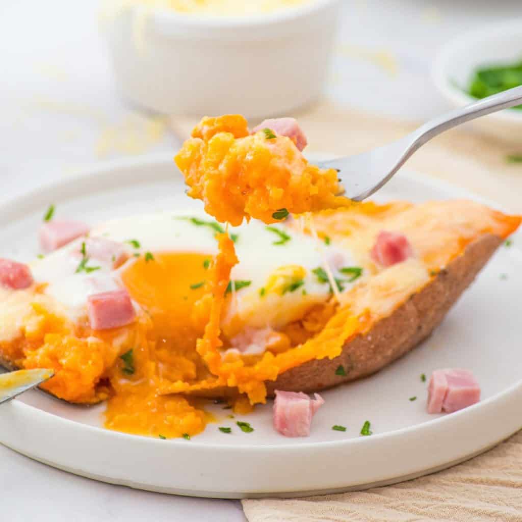 A baked sweet potato topped with a runny egg, melted cheese, diced ham, and chopped herbs is served on a white plate. A fork lifts a portion of the filling.