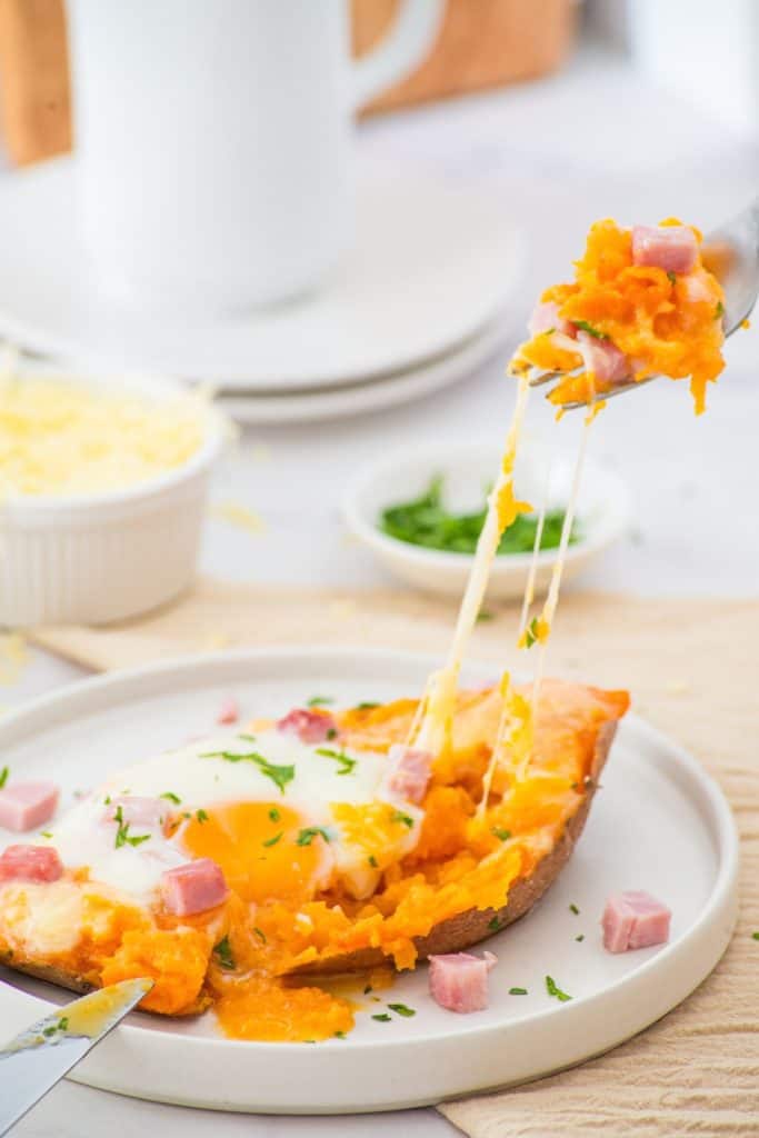 A baked sweet potato half topped with melted cheese, diced ham, and a baked egg is on a plate, with a fork lifting a cheesy bite. Small dishes of shredded cheese and chopped herbs are in the background.
