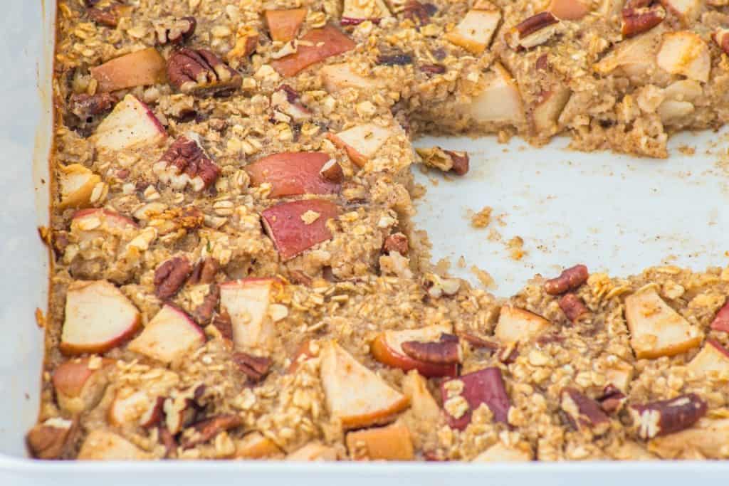 A close-up of a baked oatmeal dish with chopped apples and pecans in a white baking pan. A large square section has been cut out and removed, revealing the inside texture.