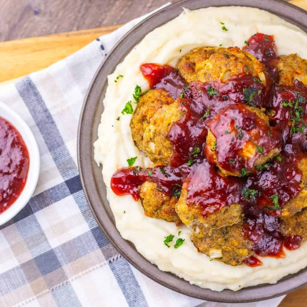 A plate of mashed potatoes topped with turkey stuffing meatballs and cranberry sauce, garnished with herbs. A small bowl of extra sauce is on the side, all beautifully plated atop a plaid cloth.