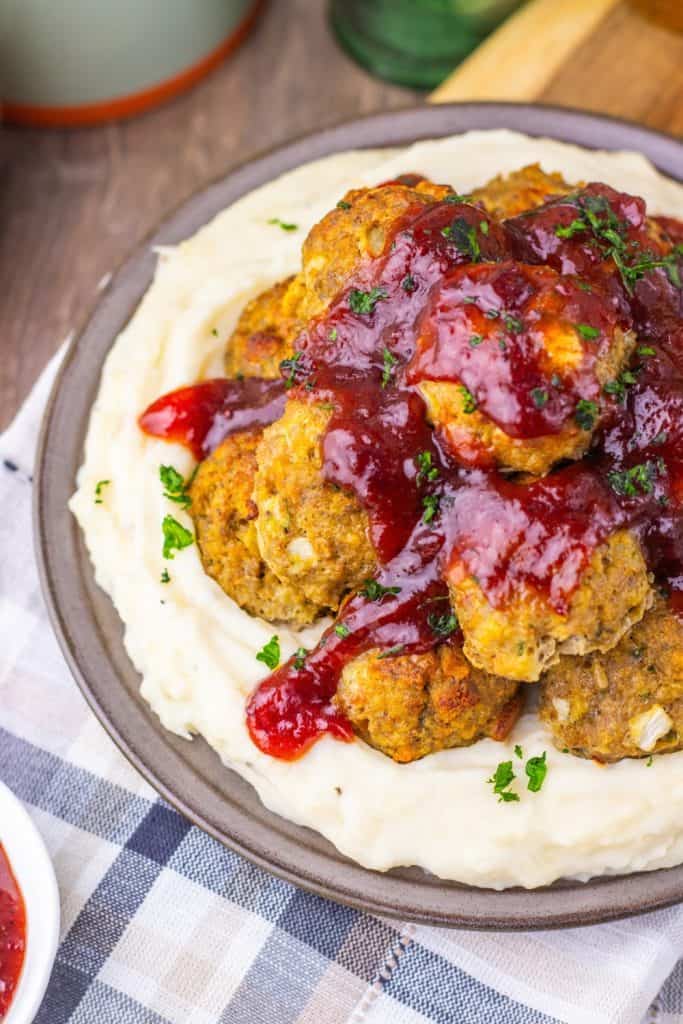 A plate of Swedish meatballs is served on mashed potatoes, topped with a generous amount of red lingonberry sauce and garnished with chopped parsley.