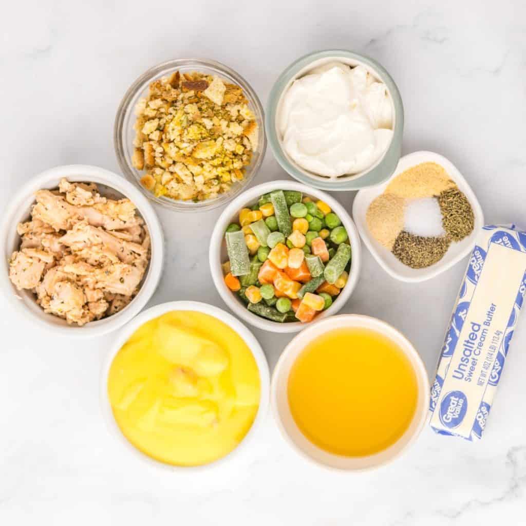 Seven bowls and a stick of butter on a white surface display ingredients for Chicken Pot Pie Casserole with Stuffing: canned chicken, mixed vegetables, soup, crushed crackers, sour cream, broth, and assorted spices.