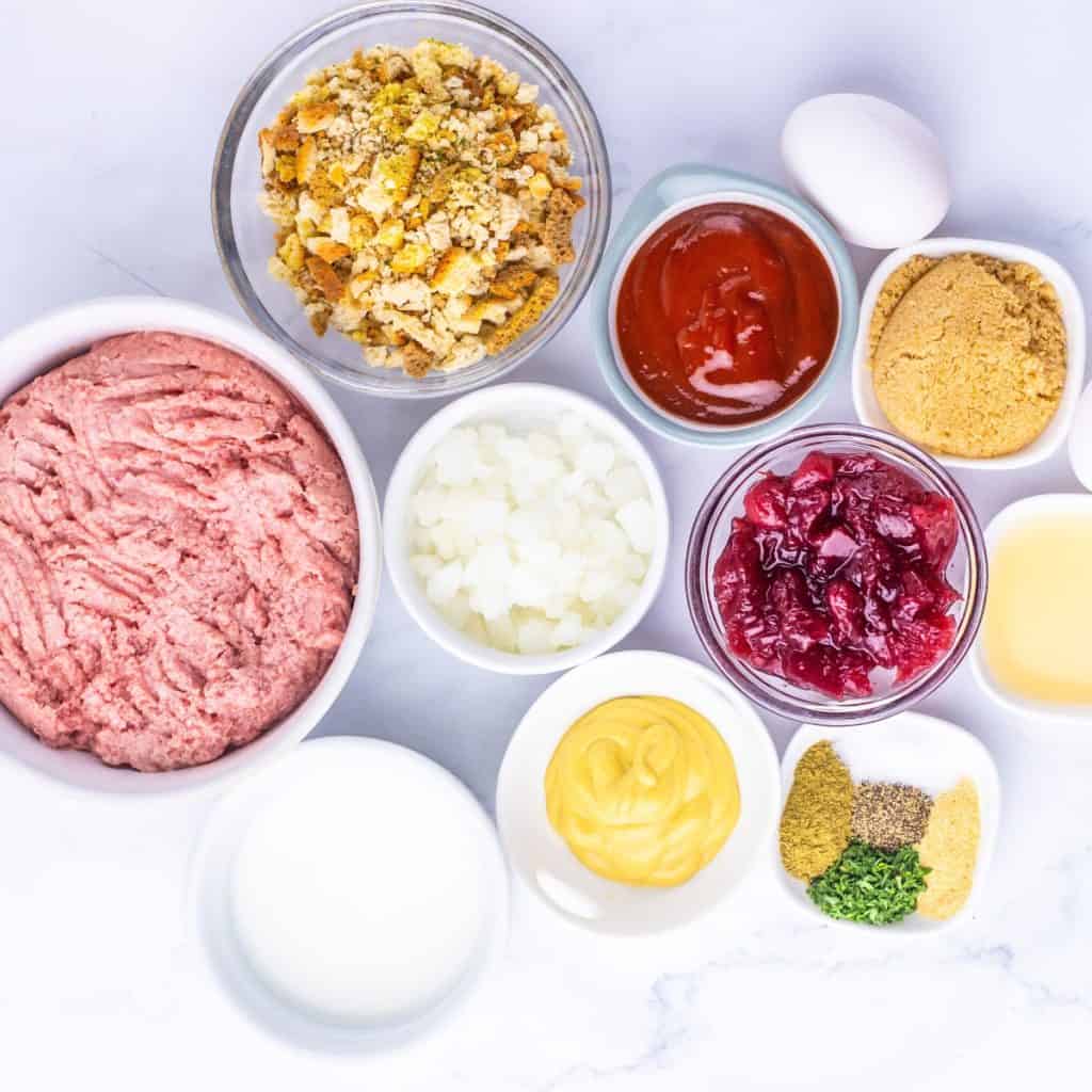 Ingredients for Turkey Stuffing Meatballs are displayed in small bowls, featuring ground meat, bread crumbs, ketchup, brown sugar, diced onion, cranberry sauce, egg, milk, mustard, apple juice, and assorted spices on a white surface.