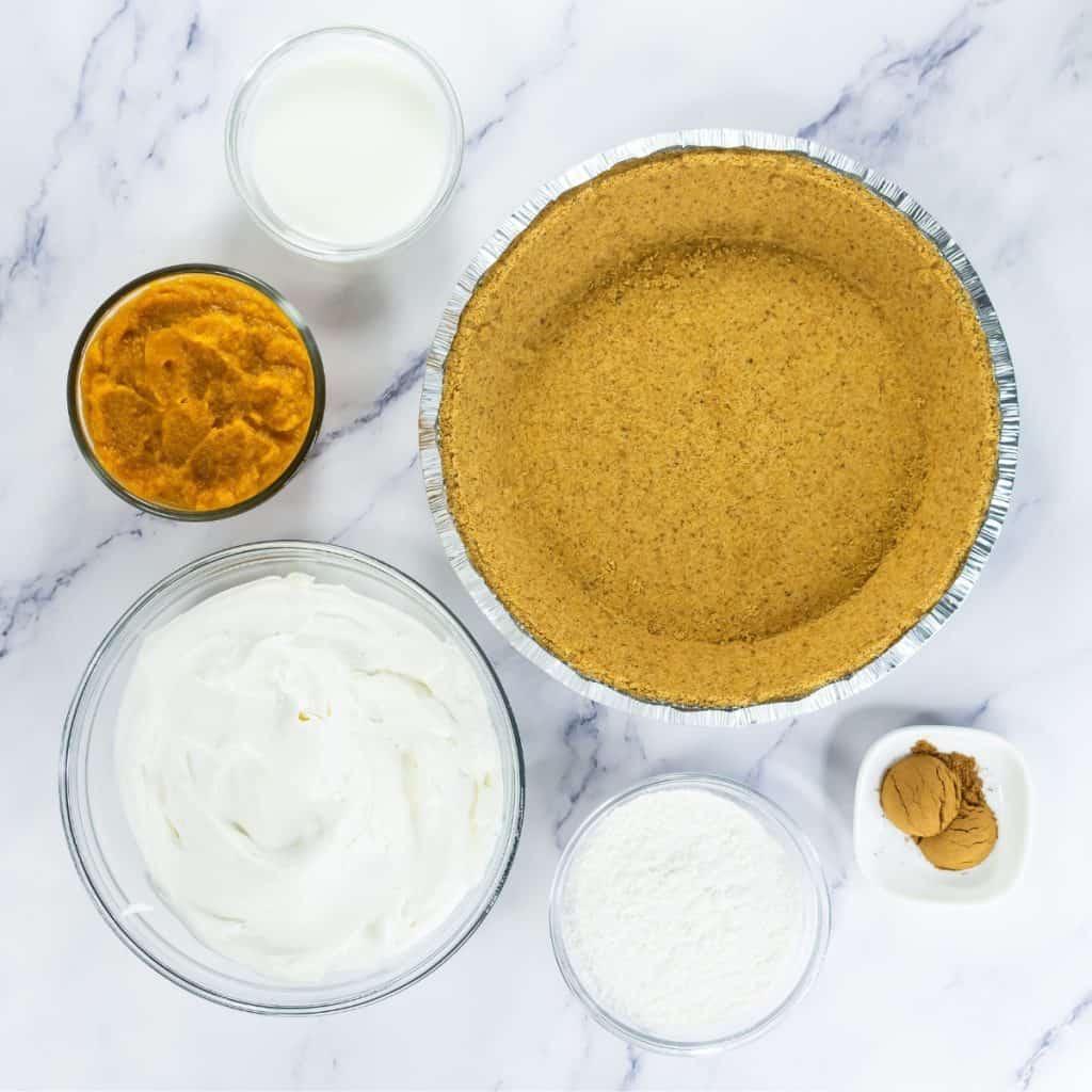 Ingredients for a no bake pumpkin pie, including a graham cracker crust, pumpkin puree, whipped topping, milk, powdered sugar, and cinnamon on a marble surface.