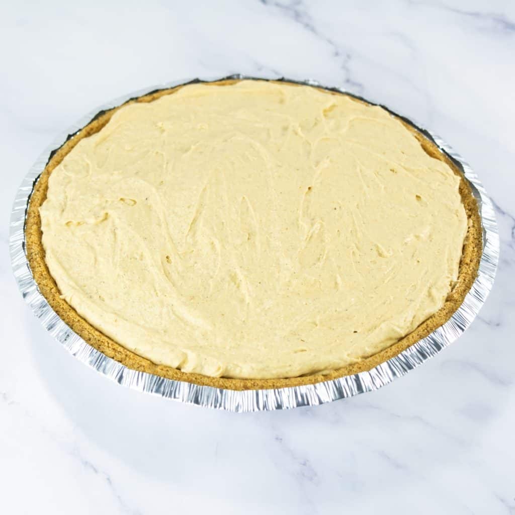 An unbaked pie with a no bake pumpkin pie with pudding creamy filling in a graham cracker crust, set in a foil pan on a white marble surface.