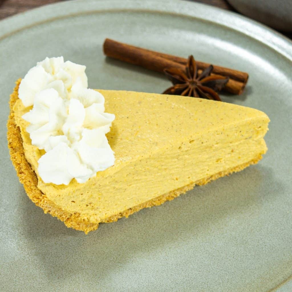 A slice of no bake pumpkin pie with vanilla pudding topped with whipped cream, served on a plate with a cinnamon stick and star anise.