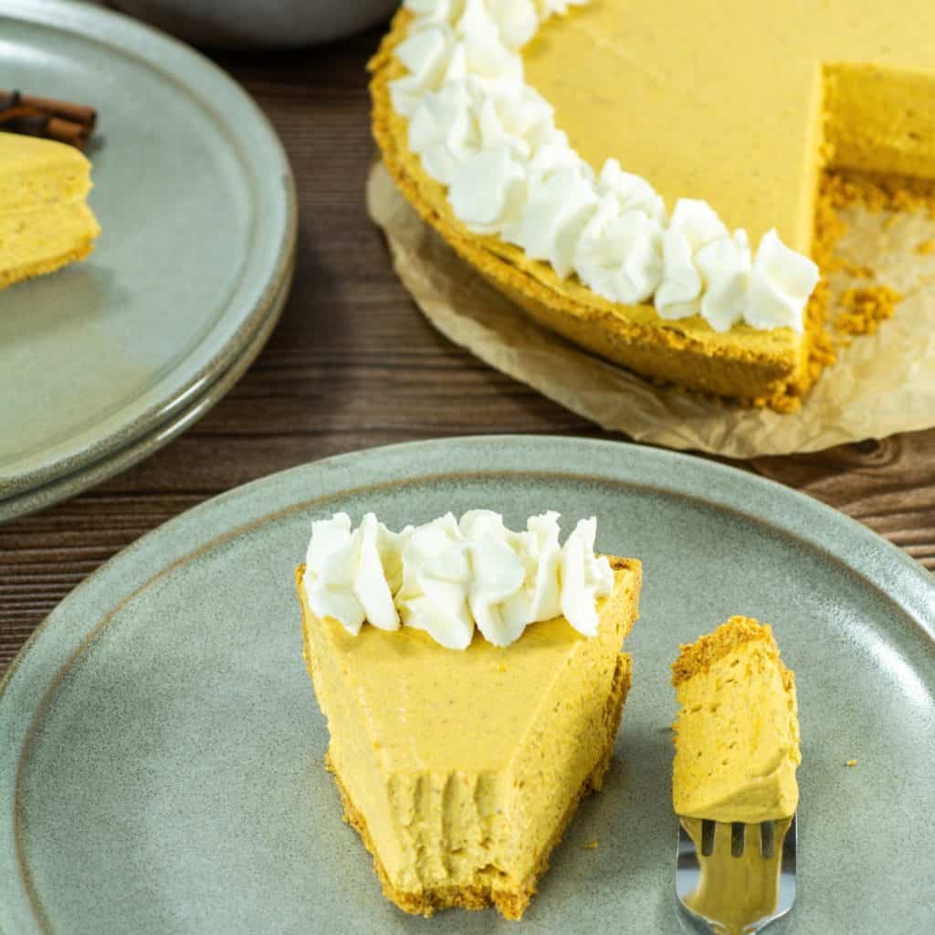 A slice of no bake pumpkin pie topped with whipped cream on a plate next to a fork, with a whole pie in the background.