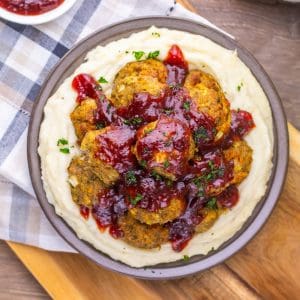 A bowl of mashed potatoes topped with several meatballs and covered in red sauce, garnished with chopped parsley.