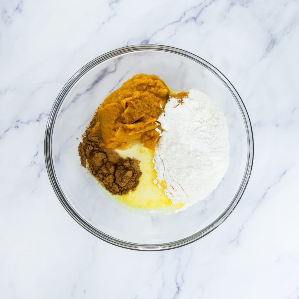 A clear glass bowl contains pumpkin purée, flour, spices, and liquid ingredients ready to be mixed for no bake pumpkin pie.
