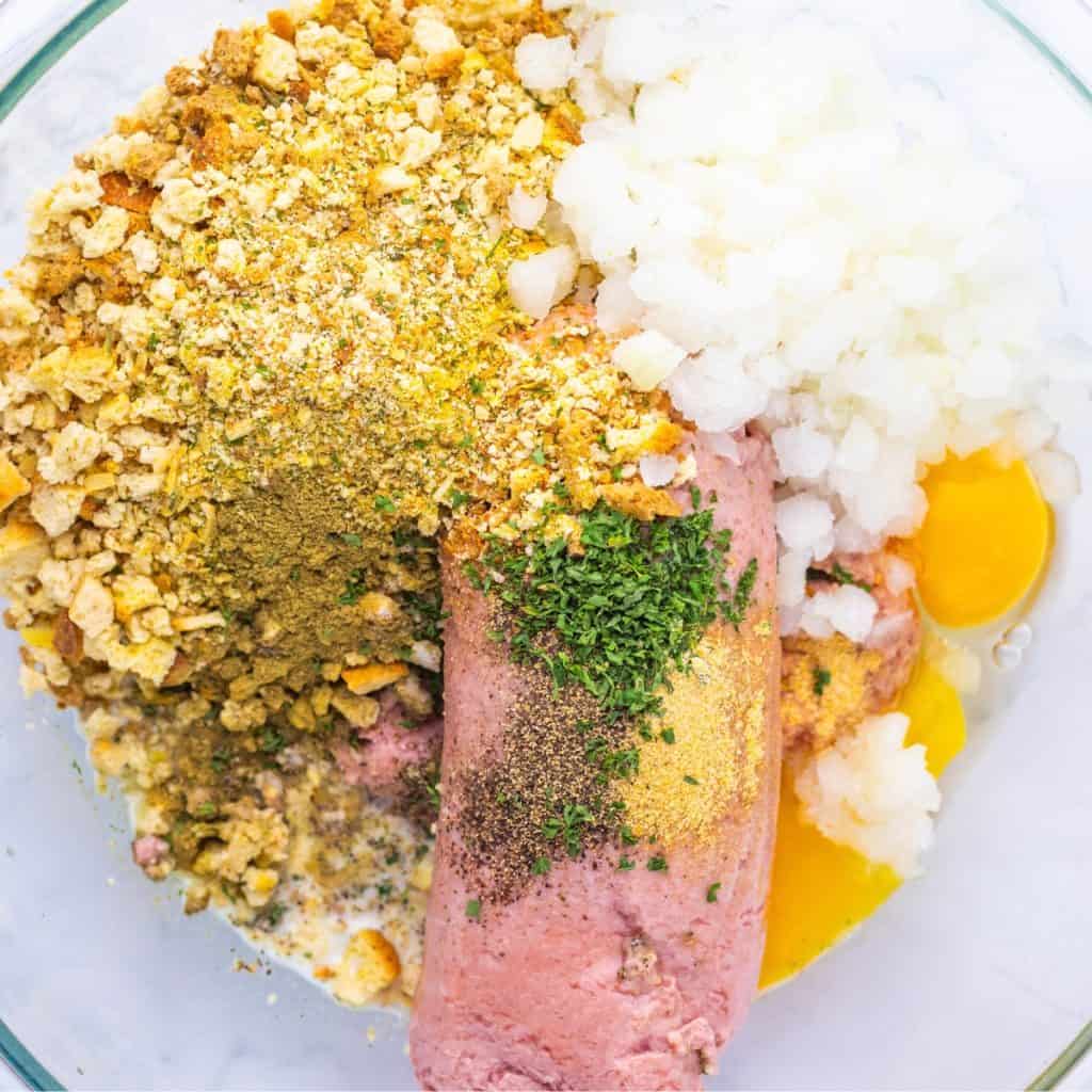 Turkey Stuffing Meatball ingredients in bowl: Ground meat, chopped onions, eggs, crushed crackers, milk, herbs, and spices are arranged in a glass mixing bowl, ready to be mixed for a flavorful dish.