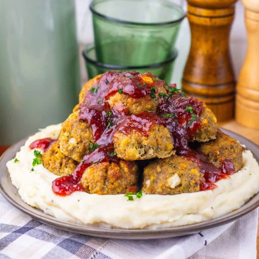 A plate of mashed potatoes topped with Turkey Stuffing Meatballs and red sauce, garnished with chopped herbs. Two stacked green glasses and salt and pepper grinders are in the background.